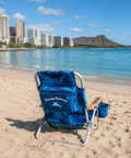 Blue beach chair with 'Tommy Bahama' branding on a sandy beach with ocean and city skyline in the background.